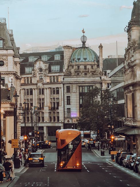 A red double-decker bus, numbered 159 with a destination sign reading 'Oxford Circus,' is parked on a busy city street with historic multi-storey buildings featuring ornate facades and large windows i