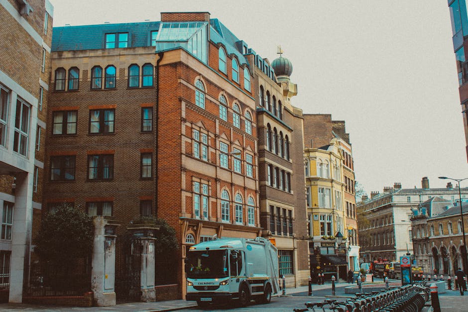 A light grey waste collection truck with a closed cargo area is parked on a city street lining a mix of historic and modern buildings. The truck, which features the logo of an independent rubbish remo